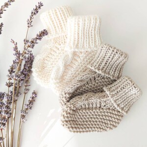 May include: Two pairs of hand-knitted baby booties in cream and light brown, with a cable knit design. The booties are displayed with sprigs of dried lavender on a white surface.