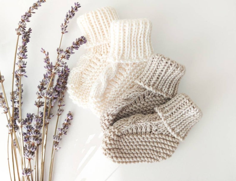 May include: Two pairs of hand-knitted baby booties in cream and light brown, with a cable knit design. The booties are displayed with sprigs of dried lavender on a white surface.