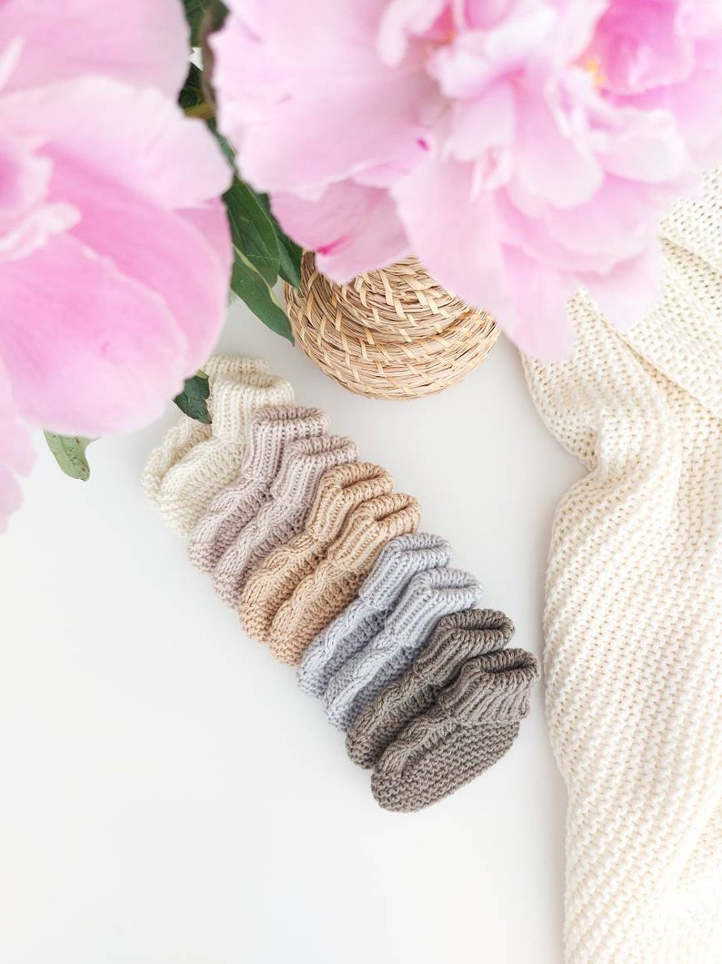 May include: A collection of seven knitted baby booties in a gradient of neutral colors, from cream to brown, arranged diagonally. The image includes pink peonies, a woven basket, and a cream-colored knitted blanket, all on a white surface.