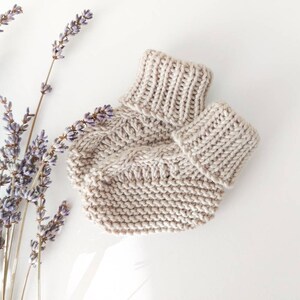 May include: A beige knitted baby hat and matching booties are displayed on a white surface, alongside sprigs of lavender. The hat features a ribbed brim and a cable knit design on the body. The booties have a similar ribbed cuff.