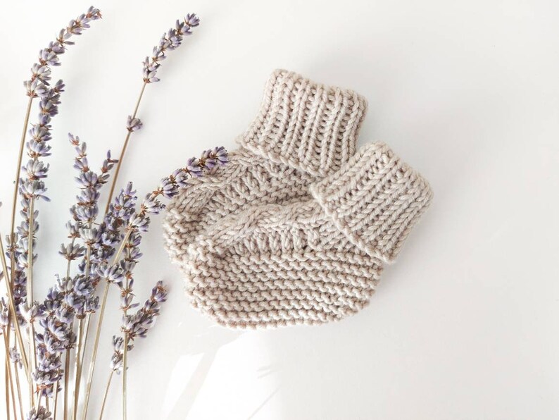 May include: A beige knitted baby hat and matching booties are displayed on a white surface, alongside sprigs of lavender. The hat features a ribbed brim and a cable knit design on the body. The booties have a similar ribbed cuff.