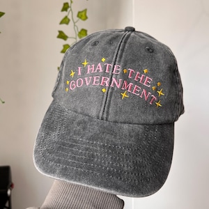 Peut inclure: Casquette de baseball grise avec l'inscription brodée en rose "I HATE THE GOVERNMENT", ornée d'étoiles jaunes. La casquette a une visière incurvée et un aspect vintage délavé.