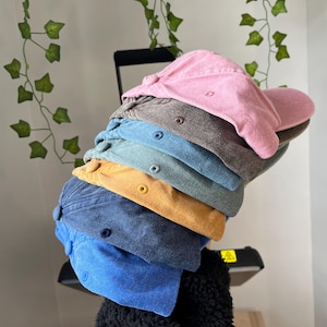 Op de afbeelding: Stapel van zes baseballpetten in verschillende kleuren, waaronder roze, bruin, teal, geel, blauw en lichtblauw. De petten zijn gemaakt van een zachte, textuurrijke stof.
