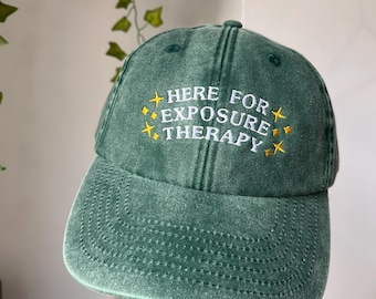 May include: Green baseball cap with the text "HERE FOR EXPOSURE THERAPY" in white and yellow. The cap has a curved brim and a distressed finish. The hat is a casual accessory.