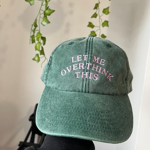 May include: A green baseball cap with the embroidered text "LET ME OVERTHINK THIS" in pink. The cap has a curved brim and a distressed look. The hat is a casual accessory.