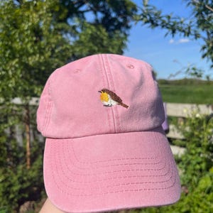 Puede incluir: Una gorra de béisbol rosa con un bordado de petirrojo en la parte delantera.