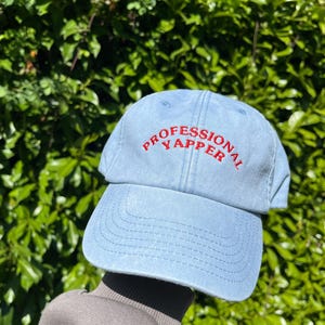 Peut inclure: Une casquette de baseball en denim bleu clair avec un texte brodé en rouge qui dit "Professional Yapper".