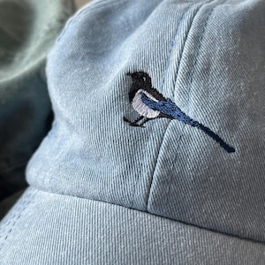 May include: A light blue denim baseball cap with an embroidered black and white magpie design.