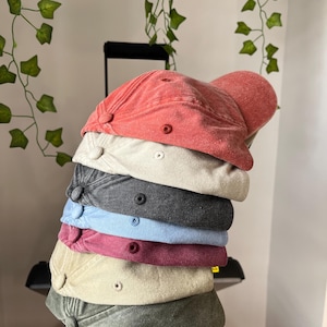 Op de afbeelding: Een stapel van acht baseballpetten in verschillende kleuren, waaronder rood, beige, zwart, blauw, bordeauxrood en groen. De petten zijn gemaakt van een zachte, gestructureerde stof.