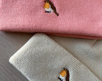 May include: Two knitted beanies with a small embroidered robin design. One beanie is pink and the other is beige.