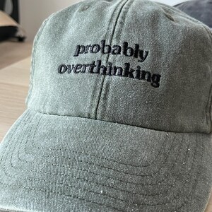 Op de afbeelding: Een groene baseballpet met zwarte geborduurde tekst die "probably overthinking" luidt.