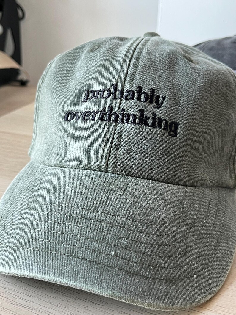 Op de afbeelding: Een groene baseballpet met zwarte geborduurde tekst die "probably overthinking" luidt.