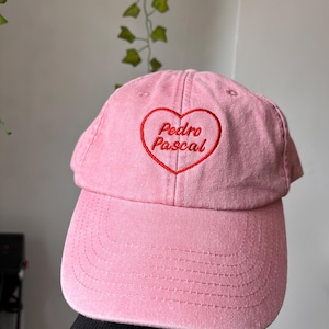 Peut inclure: Casquette de baseball rose clair avec une visière incurvée. Le nom "Pedro Pascal" est brodé en rouge à l'intérieur d'un cœur rouge. Le chapeau est en tissu doux et a un design classique à six panneaux.
