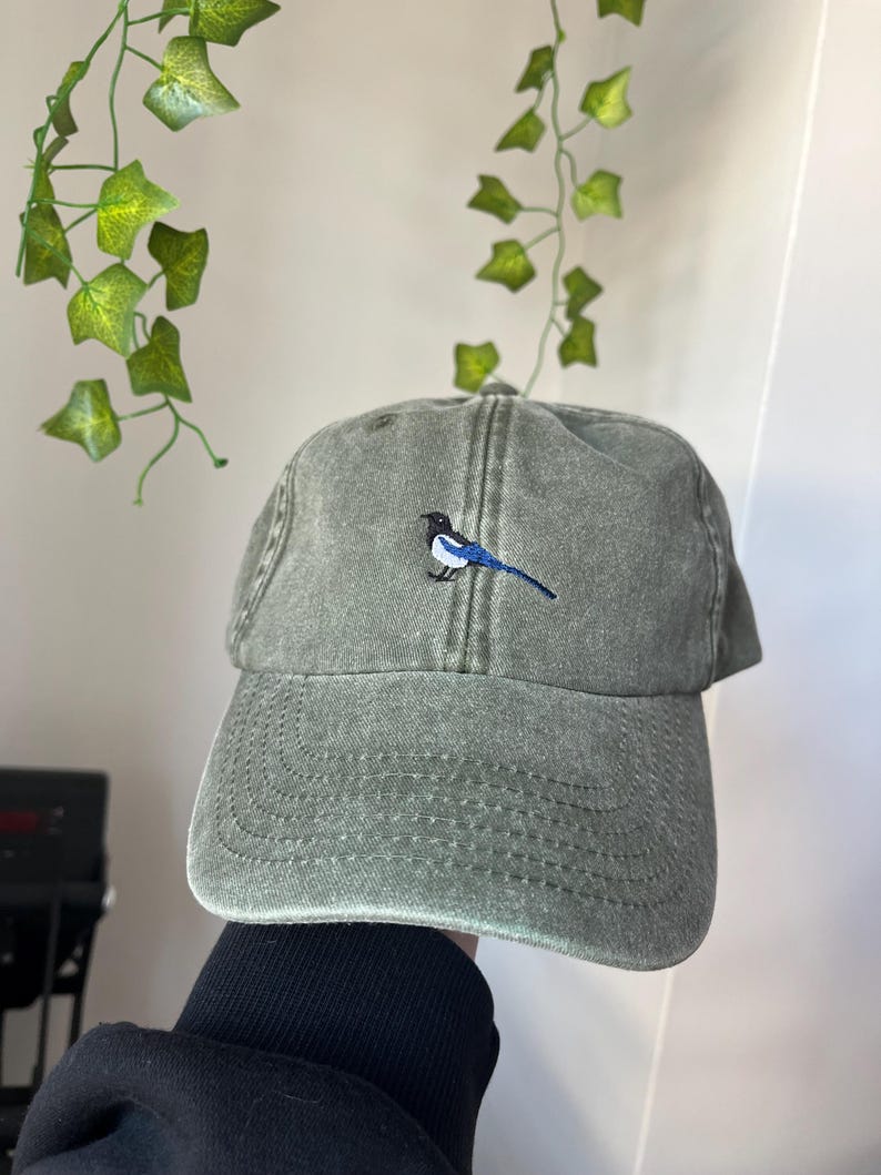 May include: A green, distressed, cotton baseball cap with an embroidered blue and black bird on the front.