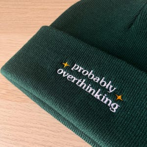 May include: A dark green beanie hat with the embroidered text "probably overthinking" in white, accented by small yellow starbursts. The hat is made of a ribbed knit fabric.
