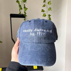 Peut inclure: Une casquette de baseball en denim bleu avec un texte brodé en blanc qui dit "easily distracted by ????".