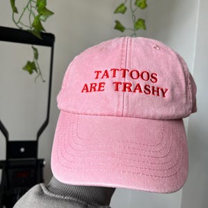 May include: A pink baseball cap with the text "Tattoos Are Trashy" embroidered in red.