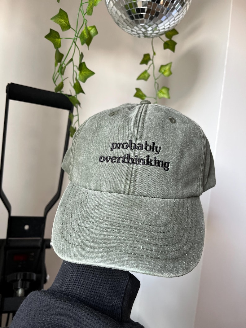 Op de afbeelding: Een groene, verweerde baseballpet met de tekst "probably overthinking" geborduurd in zwart.