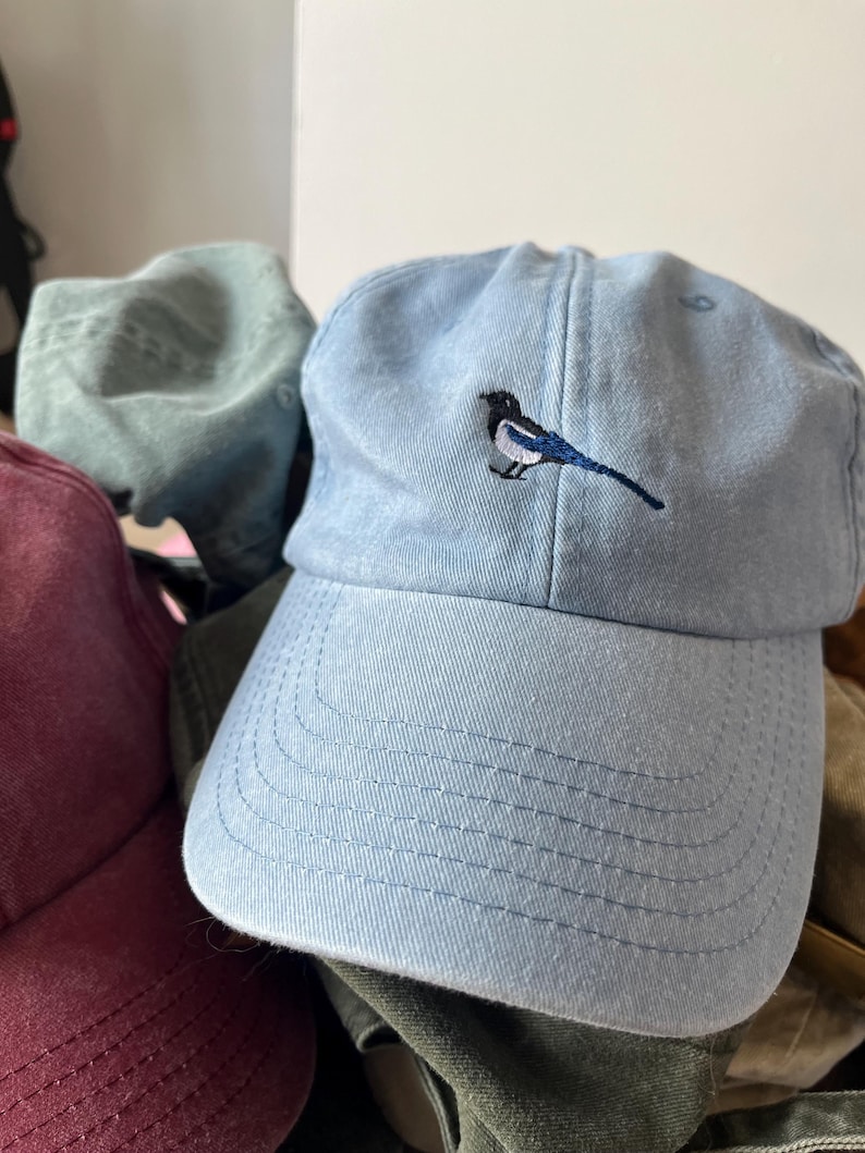 May include: A light blue baseball cap with a small embroidered design of a blue and white bird on the front.