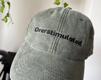 May include: A sage green baseball cap with the embroidered text "Overstimulated." The cap has a curved brim and a classic six-panel design. The hat is made of a soft, textured fabric.