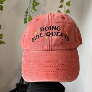 May include: A red baseball cap with the words "DOING SIDE QUESTS" embroidered in black and yellow. The cap has a curved peak and a distressed appearance. The hat is a casual accessory.