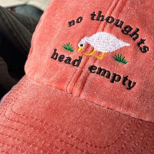 May include: A red baseball cap with the text "no thoughts head empty" embroidered on the front. A white goose with black eyes is embroidered between the words. Green grass is embroidered on either side of the goose.