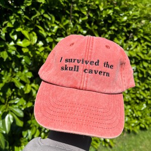 May include: A red baseball cap with the text "I survived the skull cavern" embroidered in black.