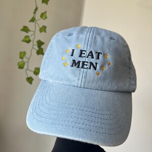 May include: Light blue baseball cap with the embroidered text "I EAT MEN" in black and gold. The cap has a curved brim and a classic six-panel design. The hat is made of a soft, textured fabric.