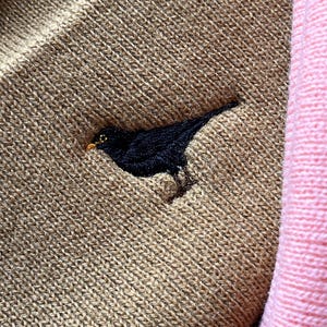 May include: A brown knitted jumper with a black embroidered bird on the front. The jumper is next to a pink knitted fabric.