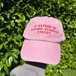 May include: A pink baseball cap with the text "I'd rather be playing Stardew Valley" embroidered in red.