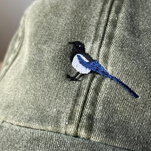 May include: A green baseball cap with an embroidered black and white magpie with blue wings.