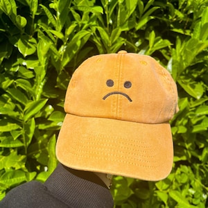 May include: Mustard-colored baseball cap with a curved brim and a stitched, adjustable back. The cap features a black embroidered sad face on the front. The hat is made of a soft, textured fabric, perfect for casual wear.