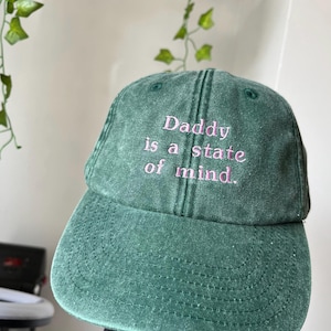 Peut inclure: Casquette de baseball verte avec le texte brodé "Daddy is a state of mind" en rose. La casquette a une visière incurvée et un design classique à six panneaux. Le chapeau est en tissu texturé.