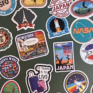 Puede incluir: Una colección de pegatinas con temática de viajes, con diseños variados. Incluyen la Torre Eiffel, un globo terráqueo y nombres de ciudades como París y Nueva York. Textos como "To Travel Is To Live" y "Enjoy your Trip" están presentes.