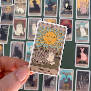 Op de afbeelding: Een tarotkaart met een witte rand en een gele zon met een gezicht in het midden. De kaart is getiteld "De Zon" en toont een grijs-witte kat met een gele halsband die in een zonnebloemenveld zit.