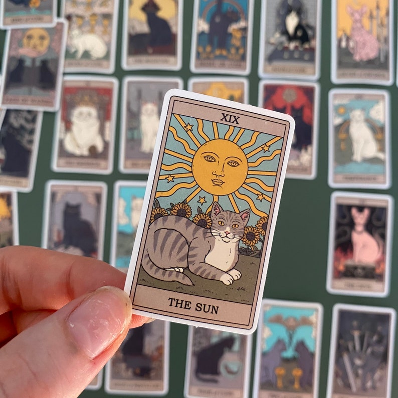 Op de afbeelding: Een tarotkaart met een witte rand en een gele zon met een gezicht in het midden. De kaart is getiteld "De Zon" en toont een grijs-witte kat met een gele halsband die in een zonnebloemenveld zit.