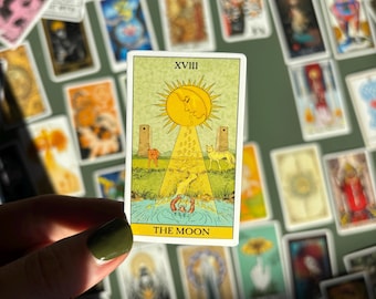 May include: A tarot card featuring a yellow sun with a crescent moon, rays of light, and the text "THE MOON". The card depicts a landscape with two towers, a dog, and a wolf. Other tarot cards are in the background.