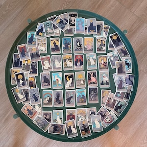 Op de afbeelding: Een set van 78 tarotkaarten met zwarte katten in verschillende poses en omgevingen. De kaarten zijn ge&iuml;llustreerd in een vintage stijl en hebben een witte achtergrond. De kaarten zijn in een cirkel op een groene ondergrond gerangschikt.