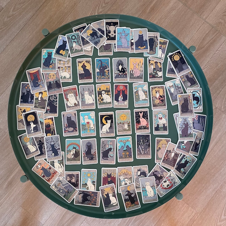 Op de afbeelding: Een set van 78 tarotkaarten met zwarte katten in verschillende poses en omgevingen. De kaarten zijn ge&iuml;llustreerd in een vintage stijl en hebben een witte achtergrond. De kaarten zijn in een cirkel op een groene ondergrond gerangschikt.