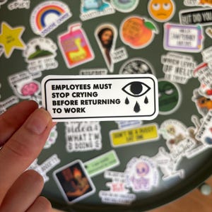 Może przedstawiać: Prostokątna biała naklejka z czarną obwódką i czarnym napisem EMPLOYEES MUST STOP CRYING BEFORE RETURNING TO WORK. Po prawej stronie tekstu znajduje się czarno-biała grafika przedstawiająca oko z trzema spadającymi łzami.
