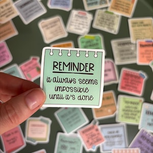 Può includere: Un adesivo quadrato color verde menta a forma di pagina di blocco note con rilegatura a spirale in alto. Il testo REMINDER è stampato in nero, seguito dalla frase it always seems impossible until it's done in un carattere corsivo scuro.