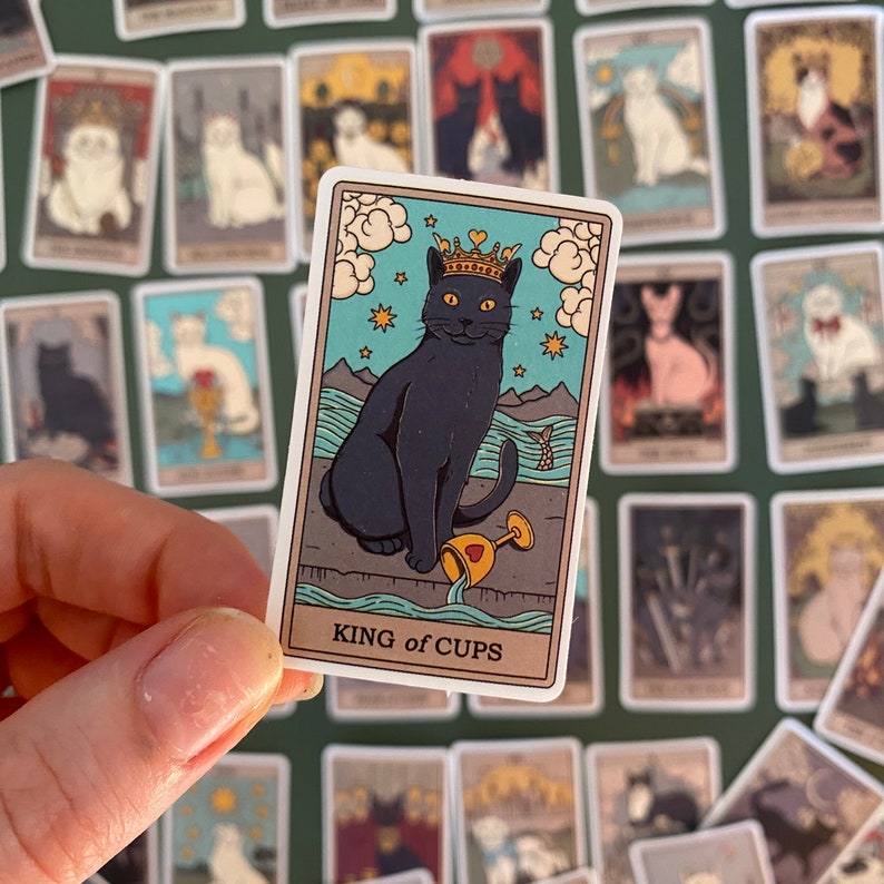 Op de afbeelding: Een zwarte kat met een gouden kroon zit op een blauwe en groene achtergrond met een gouden beker in zijn poot. De kaart toont "KING of CUPS".