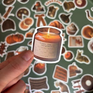 Peut inclure: Un autocollant découpé représentant une bougie en pot de verre avec une mèche allumée et une lueur orange chaude. L'étiquette porte le texte Green Tea Bergamot, scented wax candle, et divers champs administratifs. L'autocollant est tenu sur un fond.