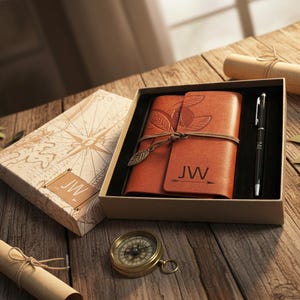Puede incluir: Un juego de diario y bolígrafo de cuero marrón en una caja de regalo. El diario tiene detalles de hojas y plumas y las iniciales "JW". También se ve una brújula y un pergamino.