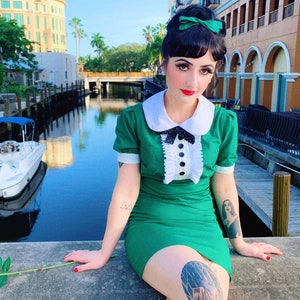 May include: A woman wearing a green dress with white collar and a black bow, sitting on a concrete wall by a canal. She has a green bow in her hair and a tattoo on her leg.