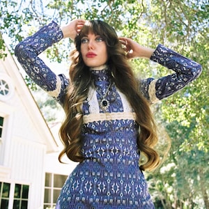 Peut inclure: Une robe bleue de style vintage avec un col montant et une garniture en dentelle blanche. La robe présente un motif détaillé dans les tons de bleu et de blanc. La personne porte un collier pendentif. L'arrière-plan comprend une maison blanche et des arbres verts.