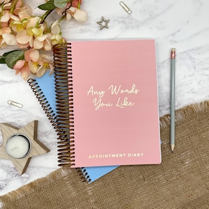 May include: A pink appointment diary with the text "Any Words You Like" in gold lettering. The diary has a brown spiral binding and is next to a grey pencil. A blue notebook is visible beneath the diary.