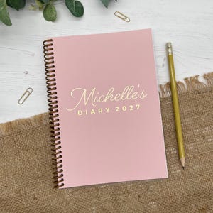 May include: A spiral-bound diary with a matte pink cover featuring the gold-coloured text Michelle's DIARY 2027. The notebook has a dark brown wire binding along the left side and is positioned next to a yellow wooden pencil with a metal ferrule.