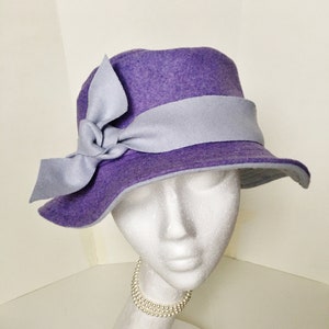 May include: A purple felt hat with a light blue felt bow and band. The hat is on a white mannequin head.