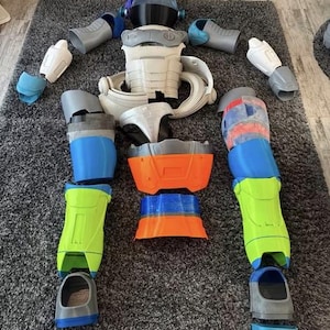 Könnte beinhalten: Ein zerlegtes Roboter-Kostüm mit verschiedenen Komponenten, die auf einem grauen Teppich angeordnet sind. Die Teile sind in Weiß, Grau, Blau, Orange und Grün gehalten. Das Kostüm umfasst einen Helm, einen Torso, Arme und Beine.